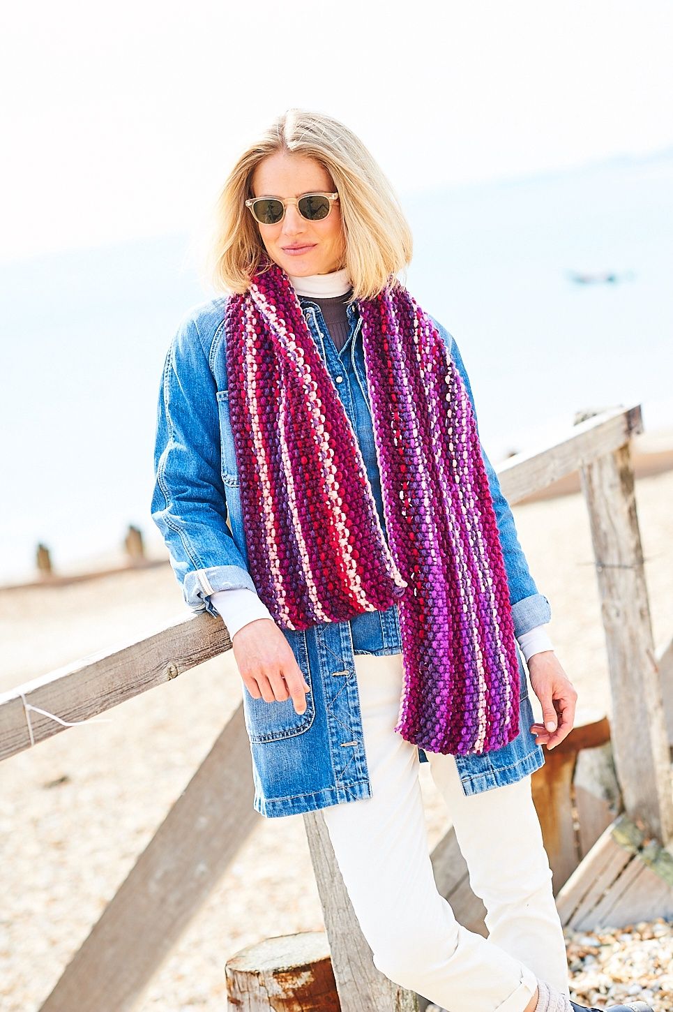 A woman is wearing a variegated knitted scarf in shades of pink, purple and red.