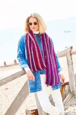 A woman is wearing a variegated knitted scarf in shades of pink, purple and red.