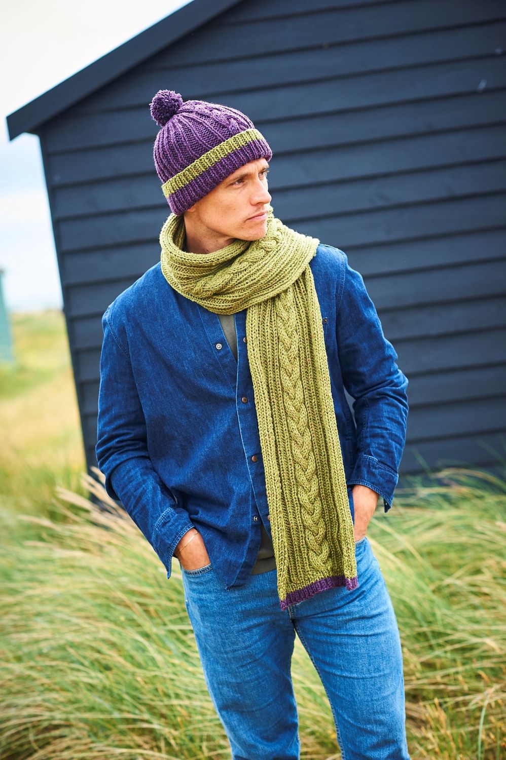 A man is standing in a field in front of a dark blue building, wearing a purple hat with a green stripe and a green scarf.