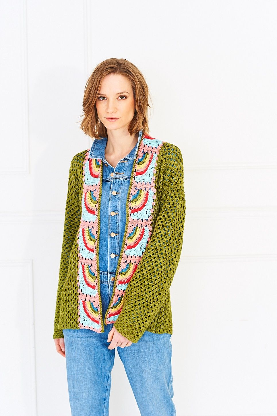 A woman is wearing a green crocheted cardigan with rainbow granny squares on the front and back.