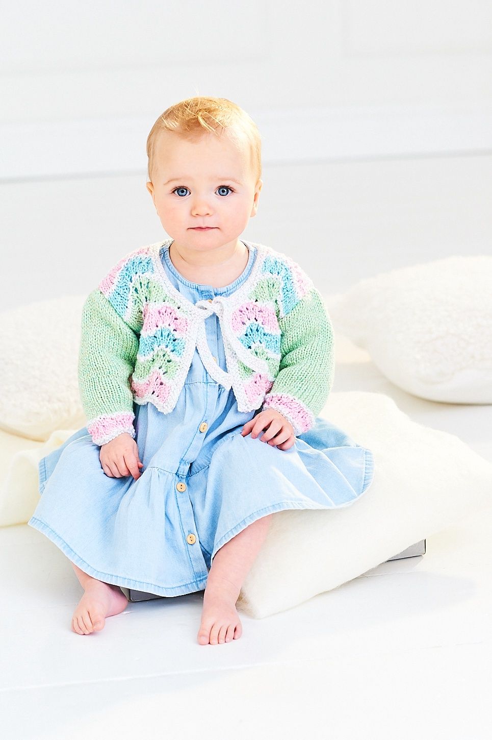 A baby is wearing a knitted ripple cardigan in green, blue, cream and pink.