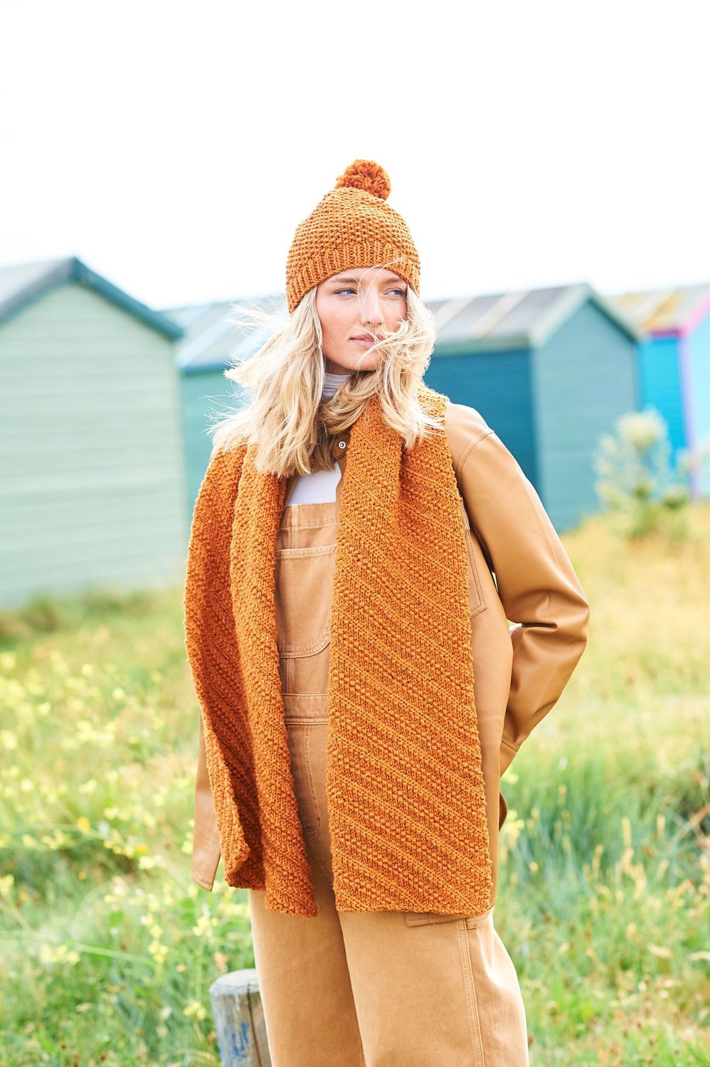 A woman is standing in a field in front of a row of blue beach huts, wearing an orange hat and scarf.
