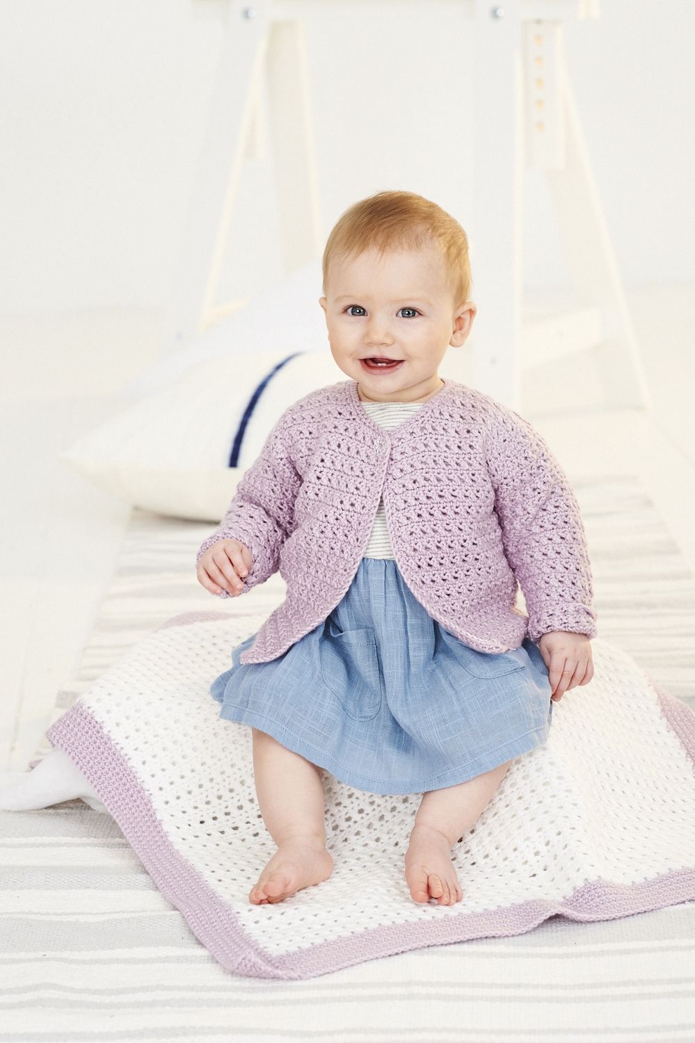 A baby is sitting on a cream crocheted blanket with a light purple border, while wearing a light purple crocheted cardigan.