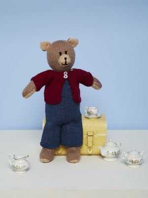 A brown knitted bear is wearing blue dungarees and a red cardigan.