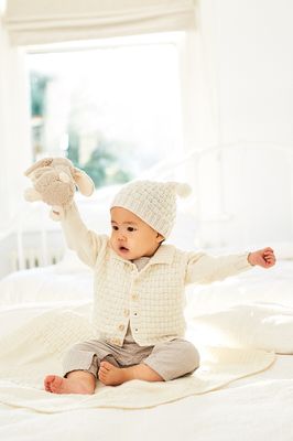 A baby is wearing a cream textured knitted cardigan and matching hat.
