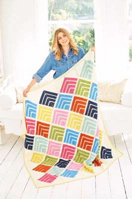 A woman is holding a crochet blanket made up of squares in cream, blue, green, pink and red.