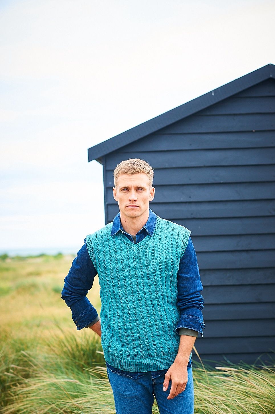 A man is stood in a field wearing a blue knitted tank top.