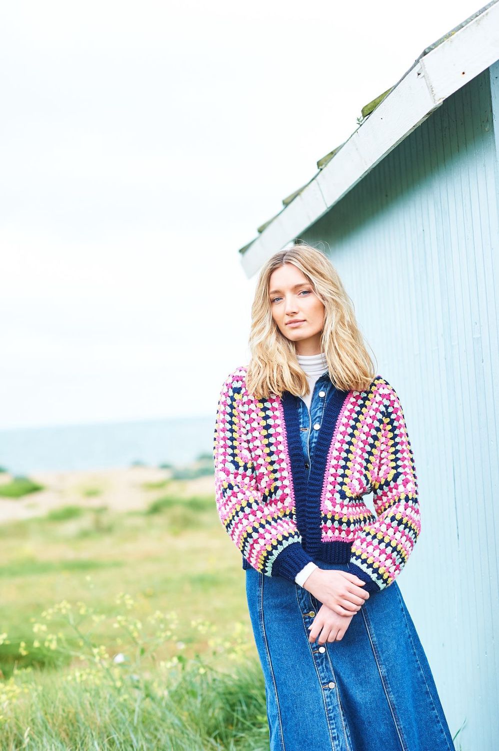 A woman is wearing a crocheted cardigan in shades of navy, pink, yellow and cream.