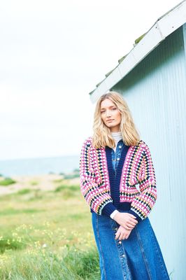 A woman is wearing a crocheted cardigan in shades of navy, pink, yellow and cream.