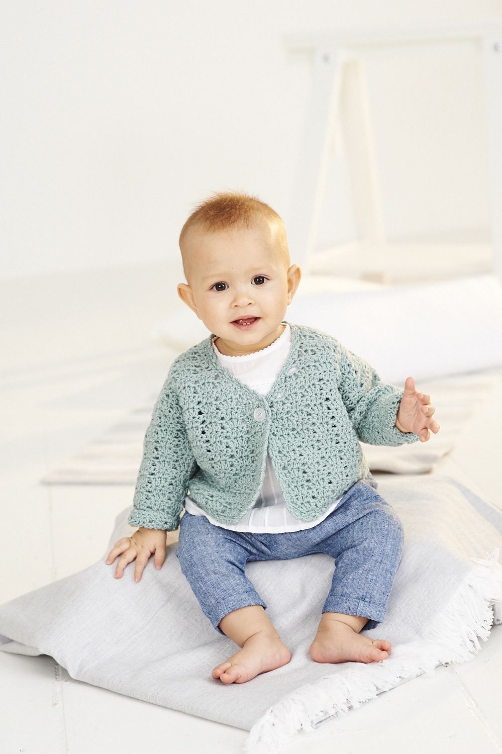 A baby is wearing a sage green crocheted cardigan.