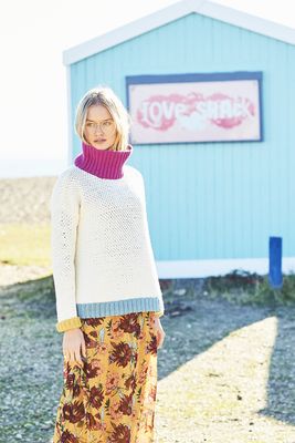 A woman is wearing a cream crocheted sweater with pink, blue and yellow cuffs.
