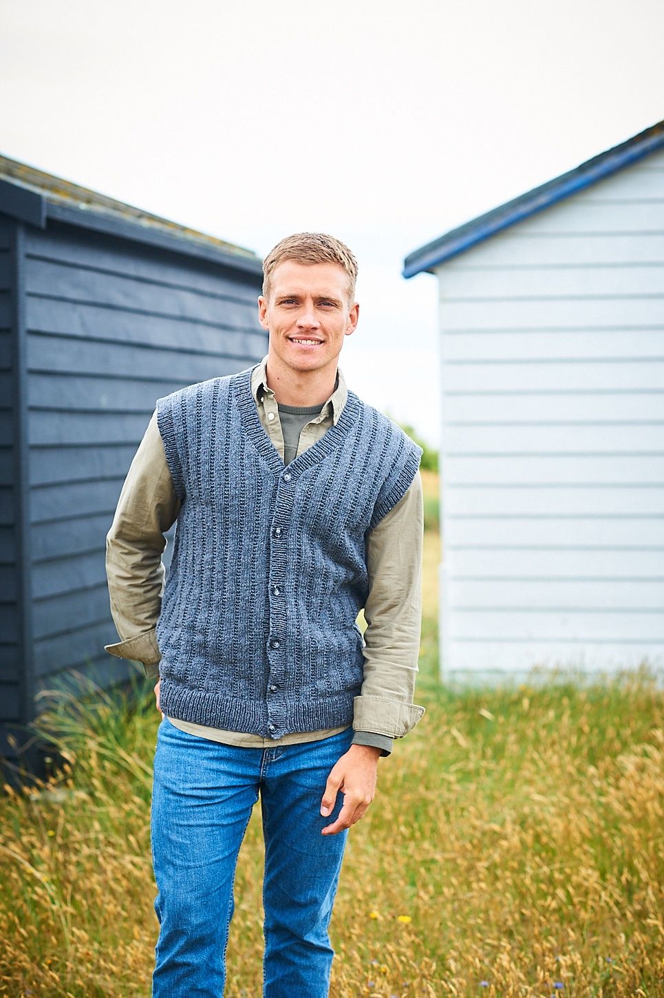 A man is stood in a field wearing a grey knitted waistcoat.