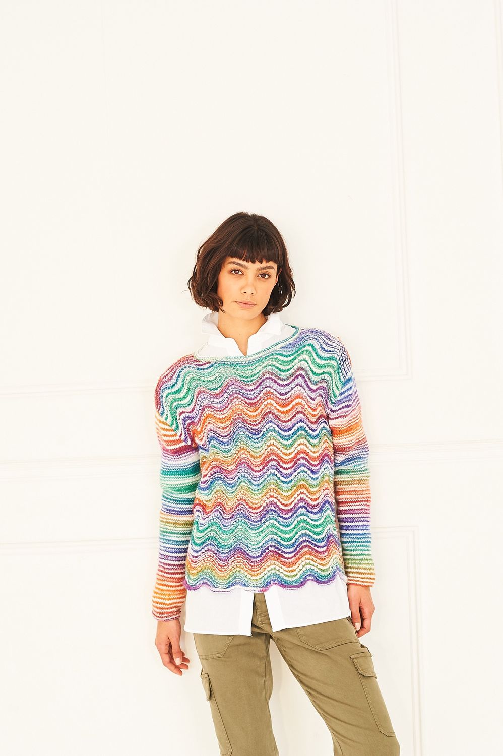A woman is wearing a knitted sweater in shades of white, red, orange, green and blue and purple.
