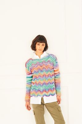 A woman is wearing a knitted sweater in shades of white, red, orange, green and blue and purple.