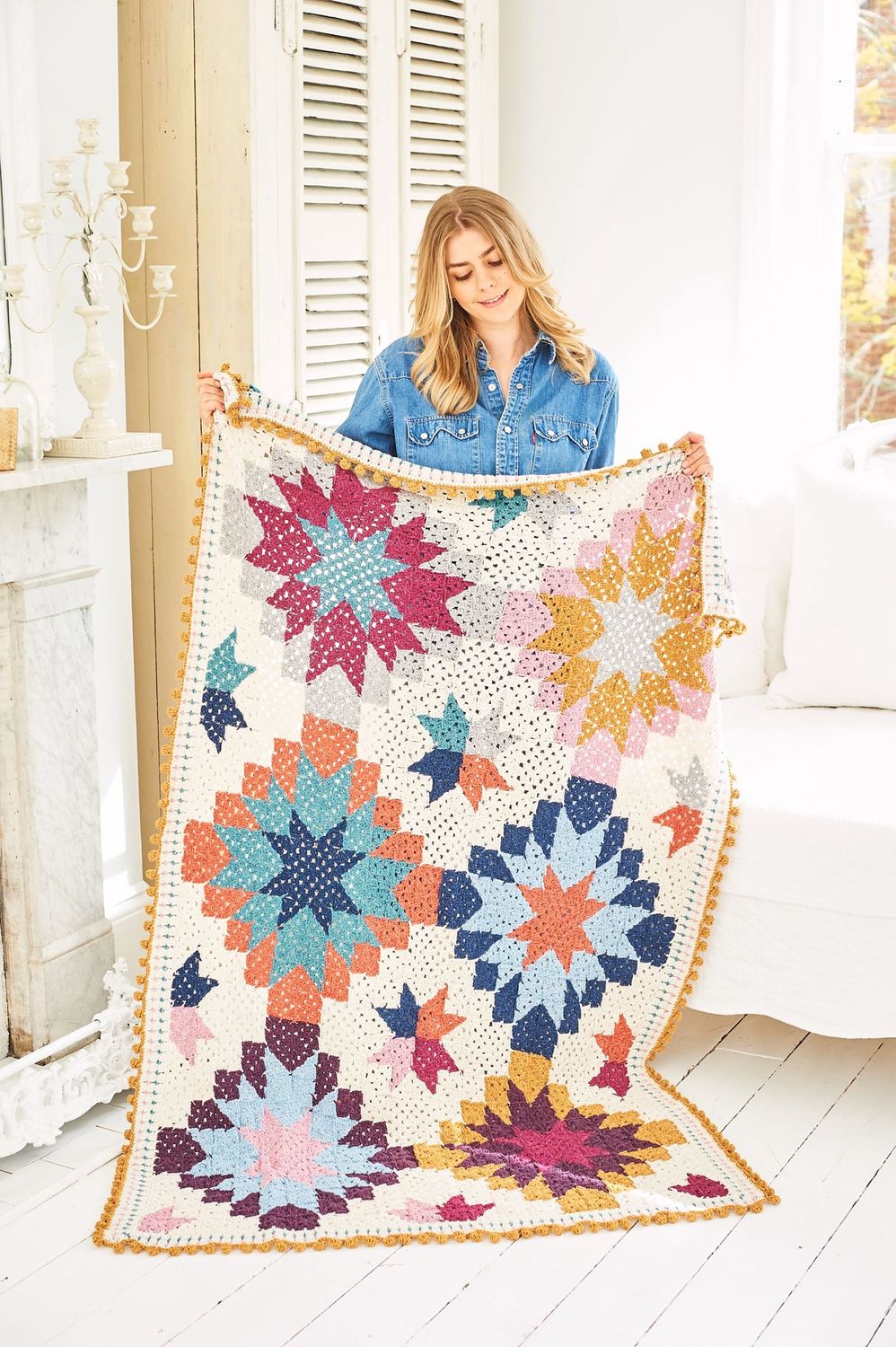 A woman is holding a crochet blanket made up of squares in cream, blue, teal, pink, yellow and silver.