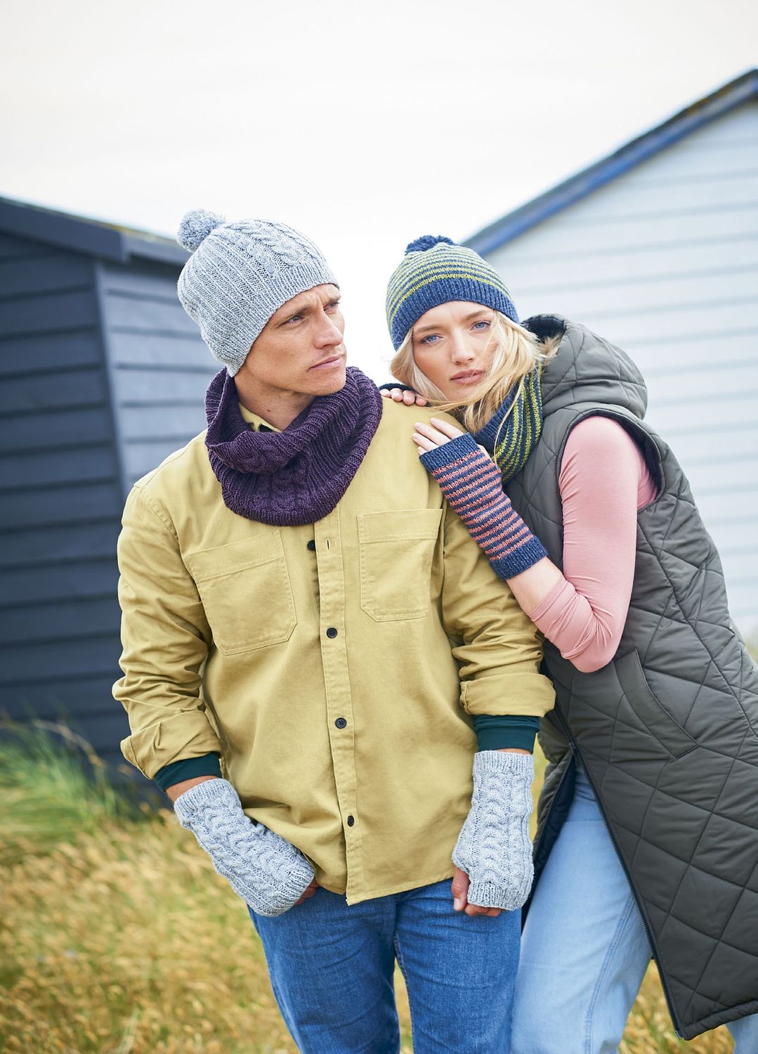 A man is wearing a grey knitted hat with matching wristwarmers and a purple knitted snood, alongside a woman who is wearing a green and blue striped knitted hat and matching snood, and pink and blue striped knitted wrsitwarmers.