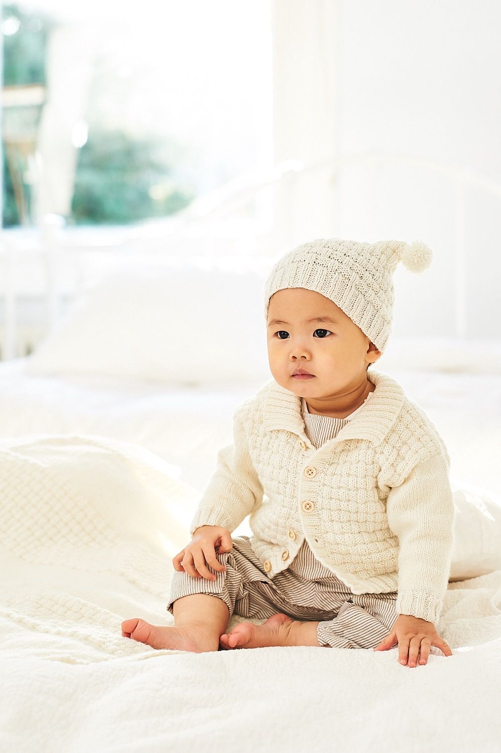 A baby is wearing a cream textured knitted cardigan and matching hat.