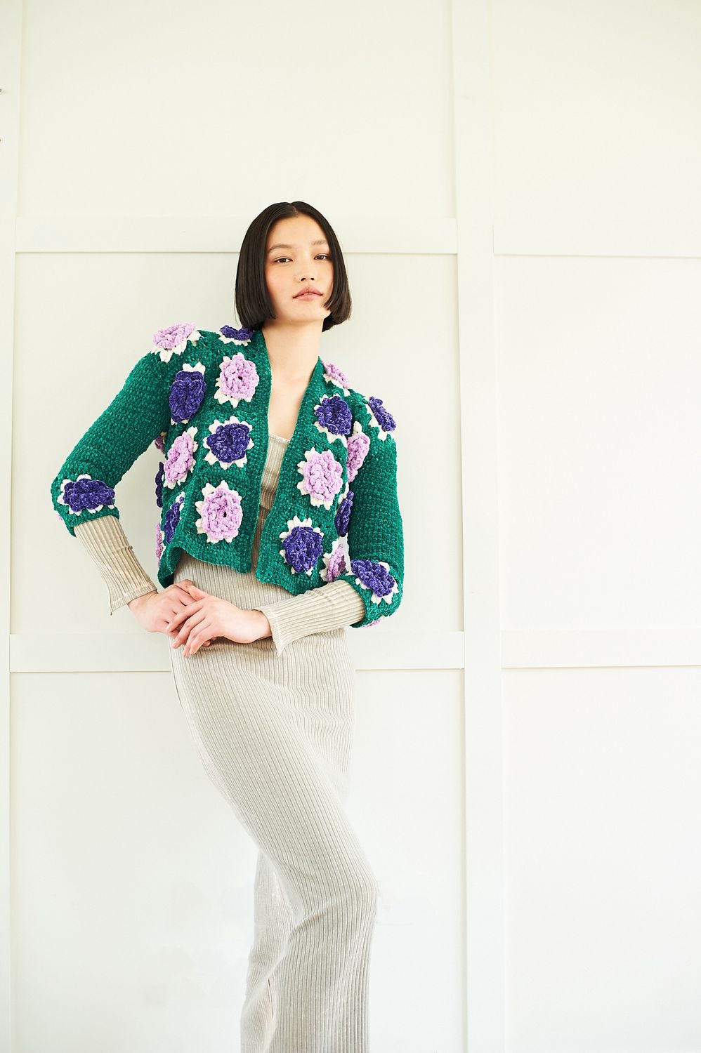 A woman is wearing a short crocheted jacket in emerald green, with purple appliqued crochet flowers.