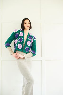 A woman is wearing a short crocheted jacket in emerald green, with purple appliqued crochet flowers.