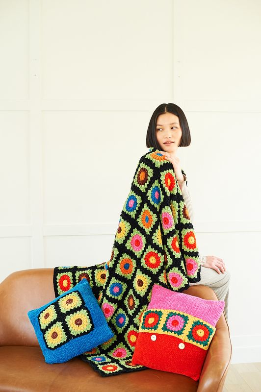 A woman is wearing a crocheted blanket in shades of black, blue, red, pink, green, yellow, brown and orange. There&#39;s also two crocheted cushions in the same shades as the blanket.