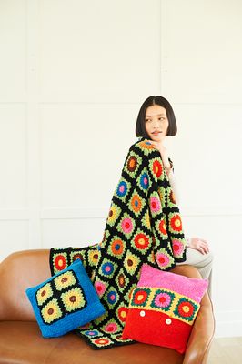 A woman is wearing a crocheted blanket in shades of black, blue, red, pink, green, yellow, brown and orange. There&#39;s also two crocheted cushions in the same shades as the blanket. 