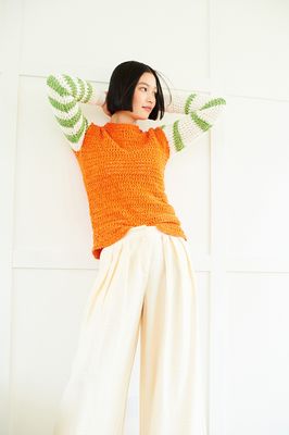 A woman is wearing an orange crocheted sweater with cream and green stripy sleeves.