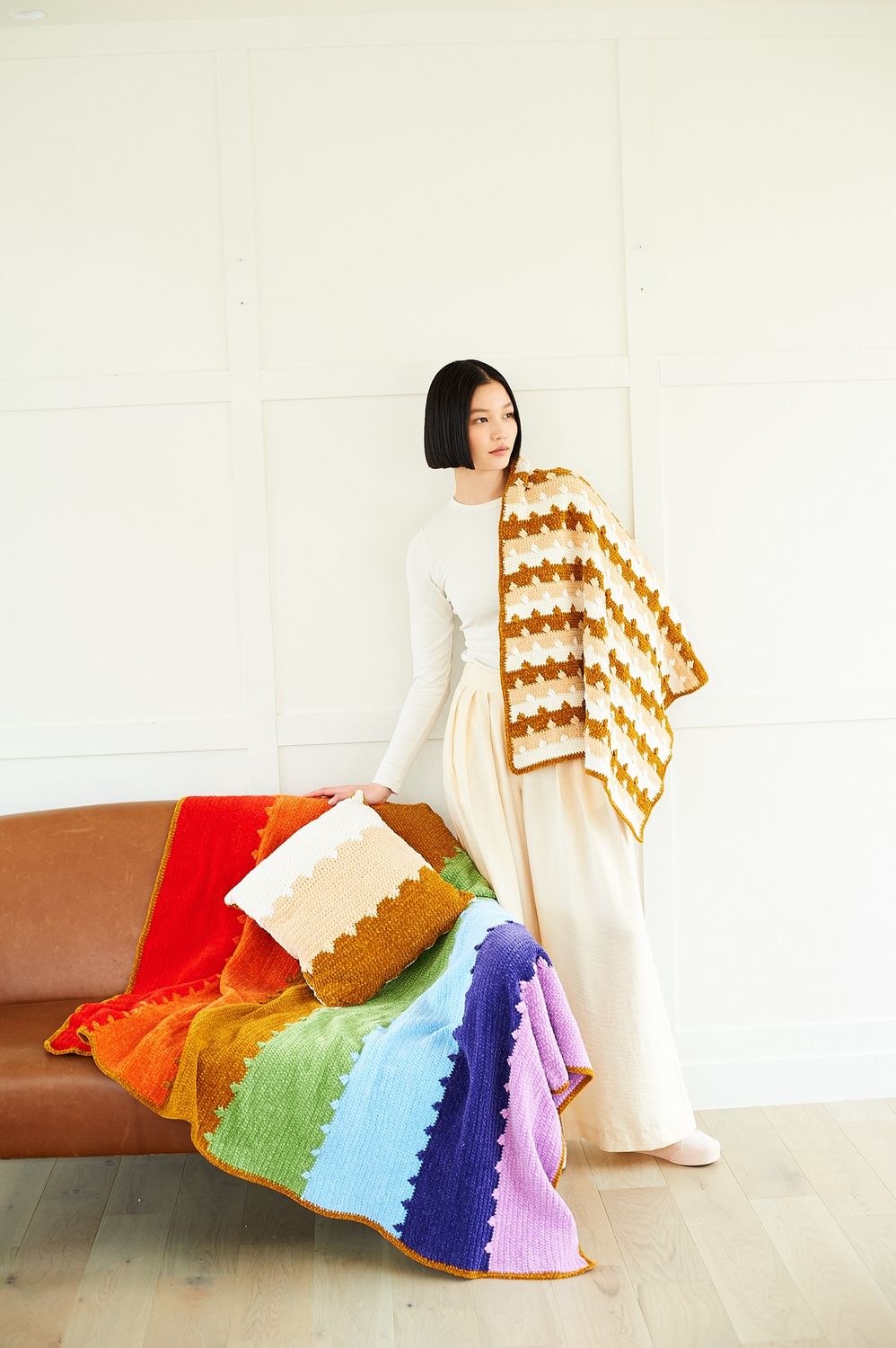 A crocheted striped blanket in shades of the rainbow and a matching crocheted striped blanket and cushion in shades of cream and brown.