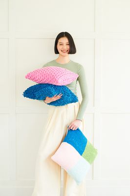 A woman is holding 3 knitted cushions, 1 blue textured, 1 pink cabled and 1 pink, green, blue and mint colourblock.