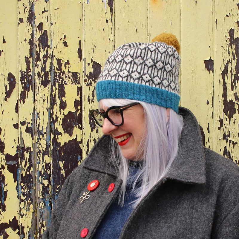 A woman is wearing a knitted colourwork hat in shades of blue, grey and cream with a mustard yellow pompom.