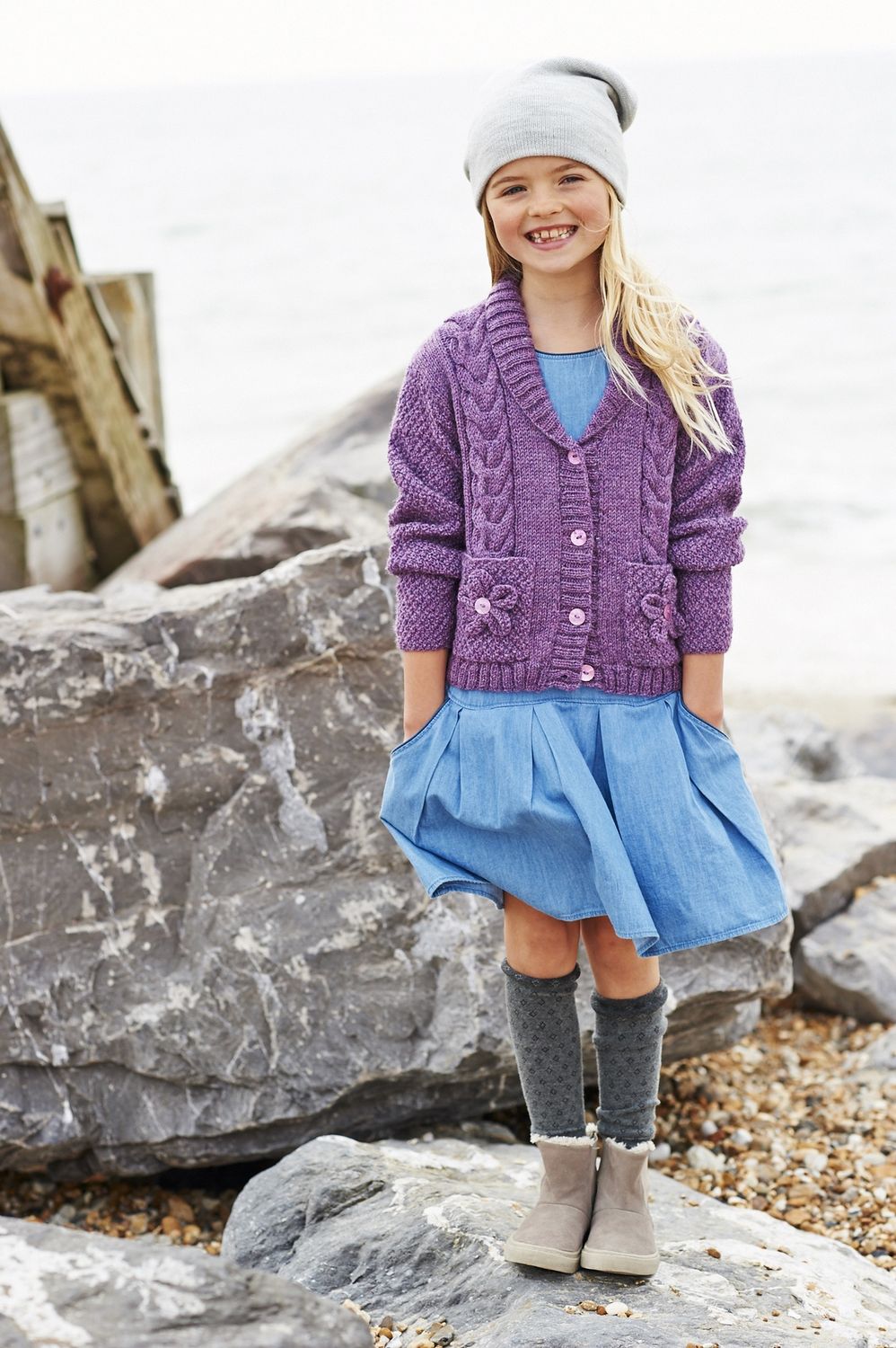 A girl is wearing a purple cable knit cardigan with flower motif pockets.