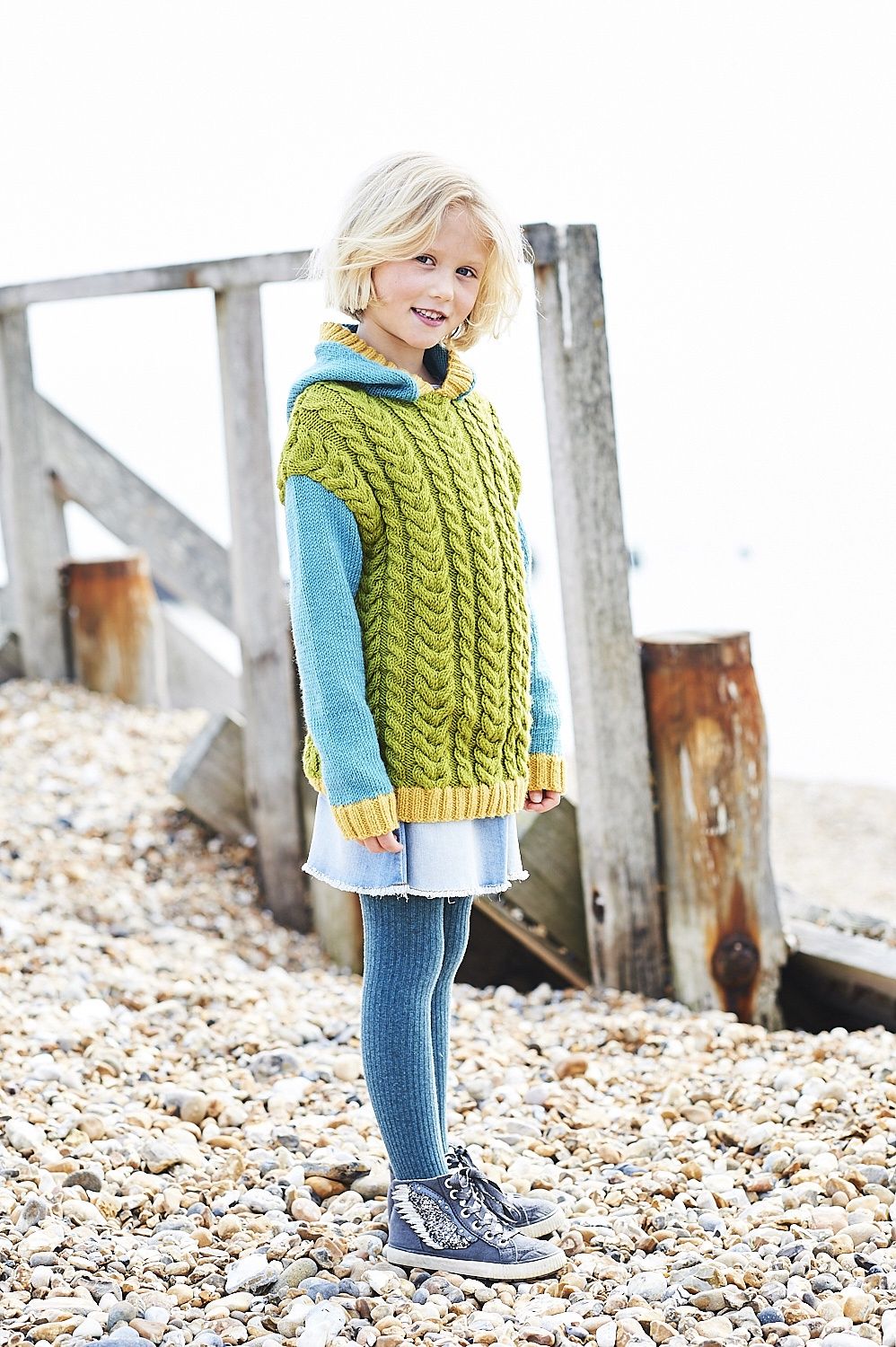 A girl is wearing a green cable knit sweater with blue sleeves and hood and yellow trim.