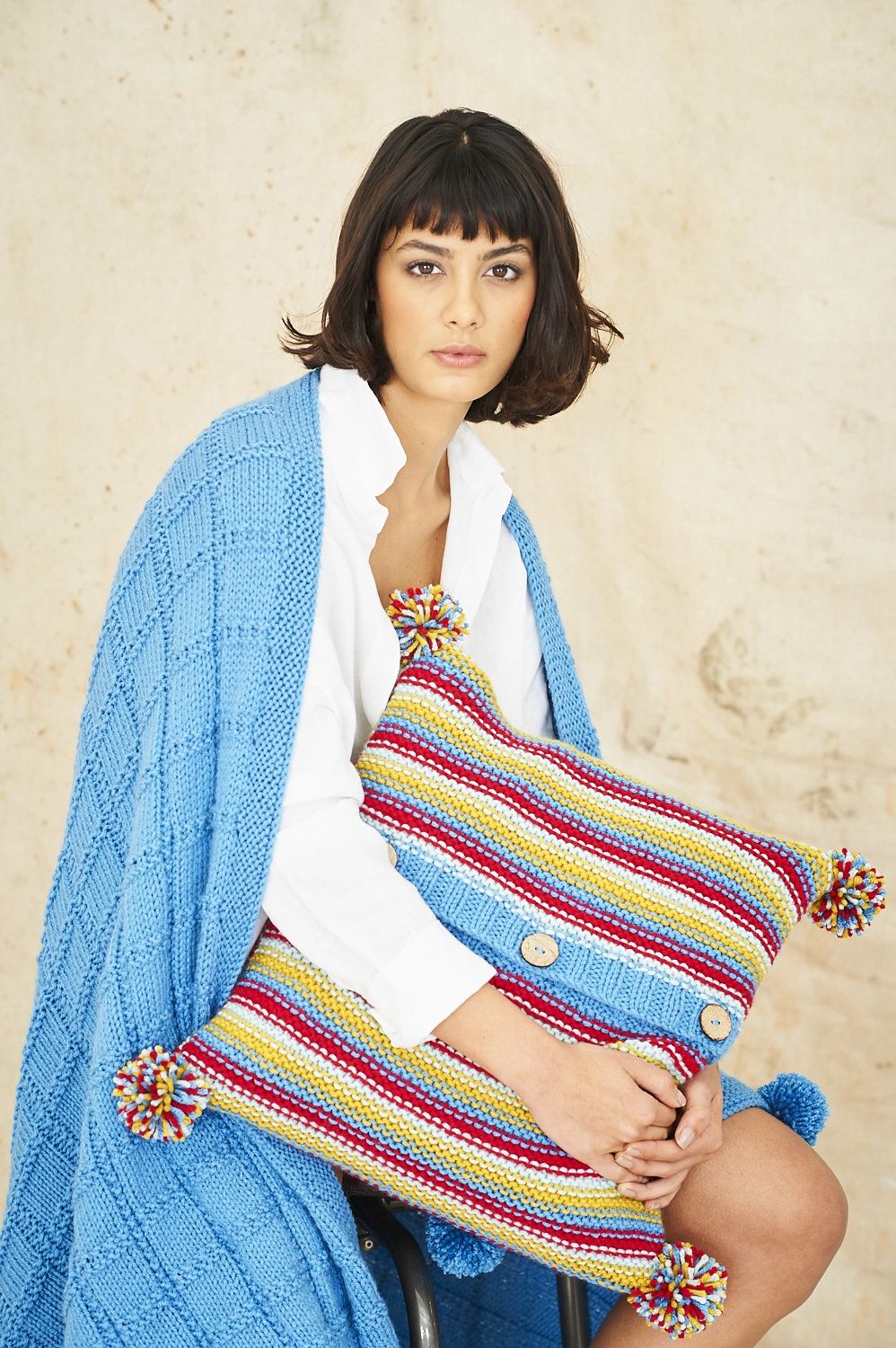 A woman is wearing a blue knitted blanket and holding a red, blue and yellow striped knitted cushion.