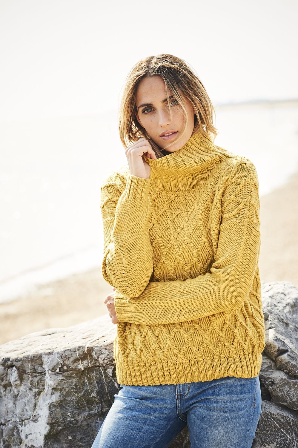 A woman is wearing a mustard yellow knitted sweater with diamond shaped cable detail.