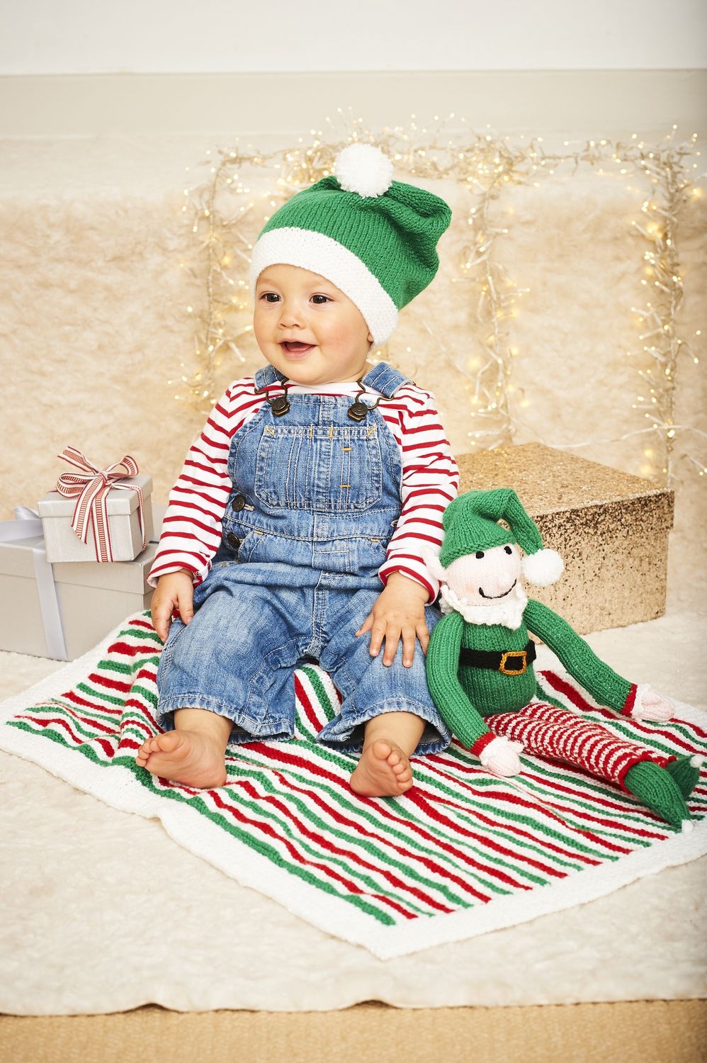 A baby is wearing a knitted green and white knitted hat, sitting on a white, green and red stripy knitted blanket with a knitted elf toy.