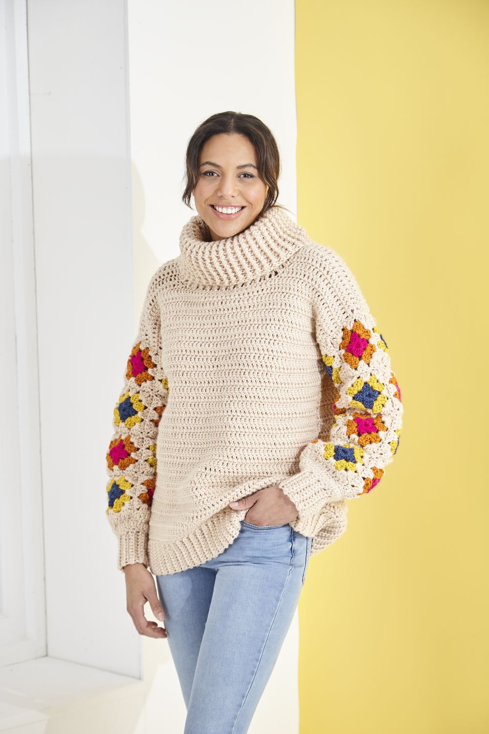 A woman is wearing a cream crocheted sweater with pink, orange, blue and yellow granny square sleeves.