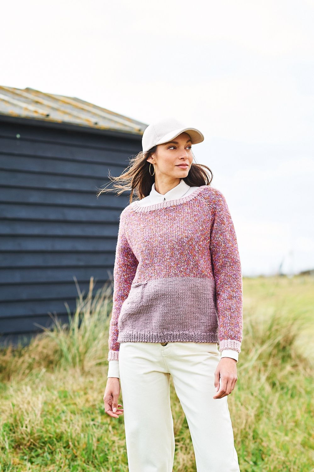 A woman is wearing a knitted sweater, with the top half in a variegated purple-brown yarn and the bottom half in solid dusky purple.