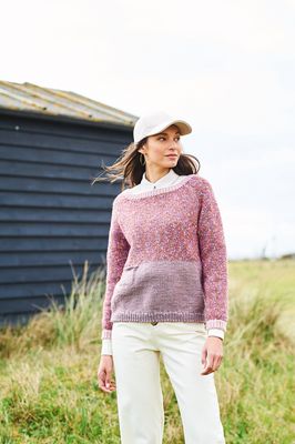 A woman is wearing a knitted sweater, with the top half in a variegated purple-brown yarn and the bottom half in solid dusky purple.