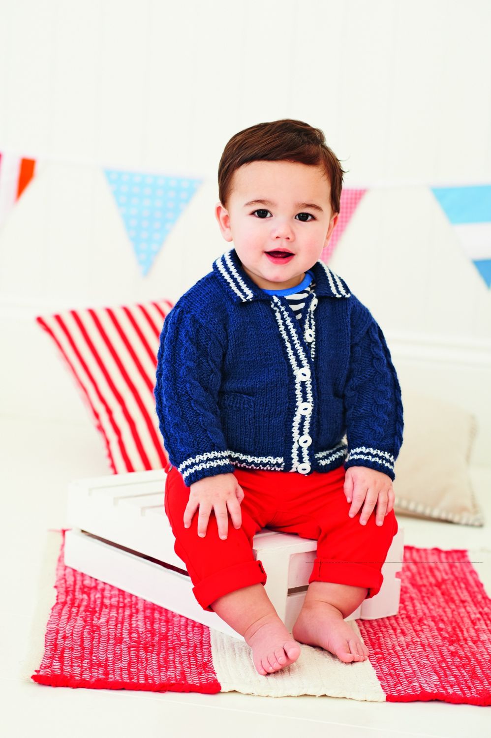 A baby is wearing a knitted cardigan in navy blue with cream edging and buttons.