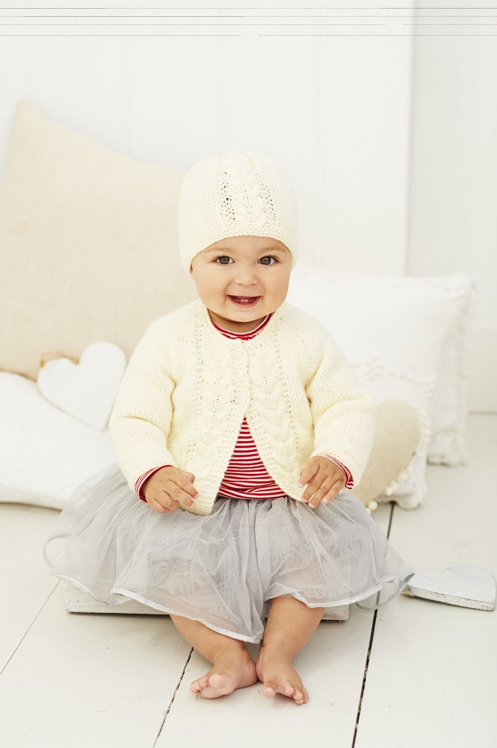 A baby is wearing a cream cable knit cardigan and matching hat.