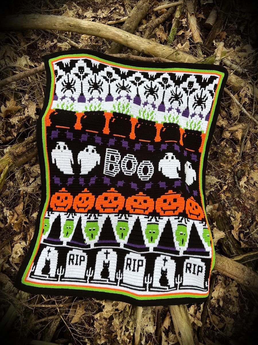 A crocheted blanket in black, white, purple, green and orange. The blanket contains various Halloween designs such as ghosts, pumpkins, spiders, zombies, cauldrons and more.