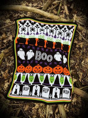 A crocheted blanket in black, white, purple, green and orange. The blanket contains various Halloween designs such as ghosts, pumpkins, spiders, zombies, cauldrons and more.