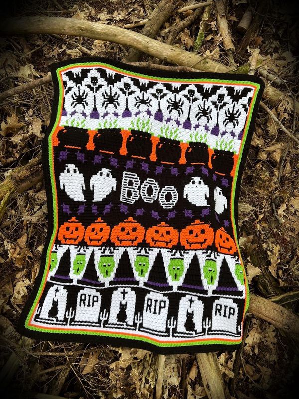 A crocheted blanket in black, white, purple, green and orange. The blanket contains various Halloween designs such as ghosts, pumpkins, spiders, zombies, cauldrons and more.