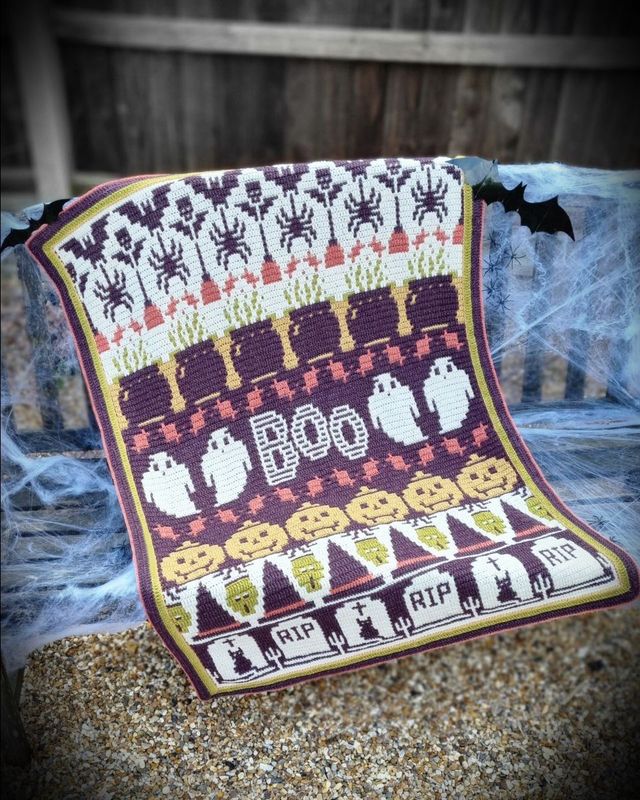 A crocheted blanket in cream, purple, red, green and yellow. The blanket contains various Halloween designs such as ghosts, pumpkins, spiders, zombies, cauldrons and more.