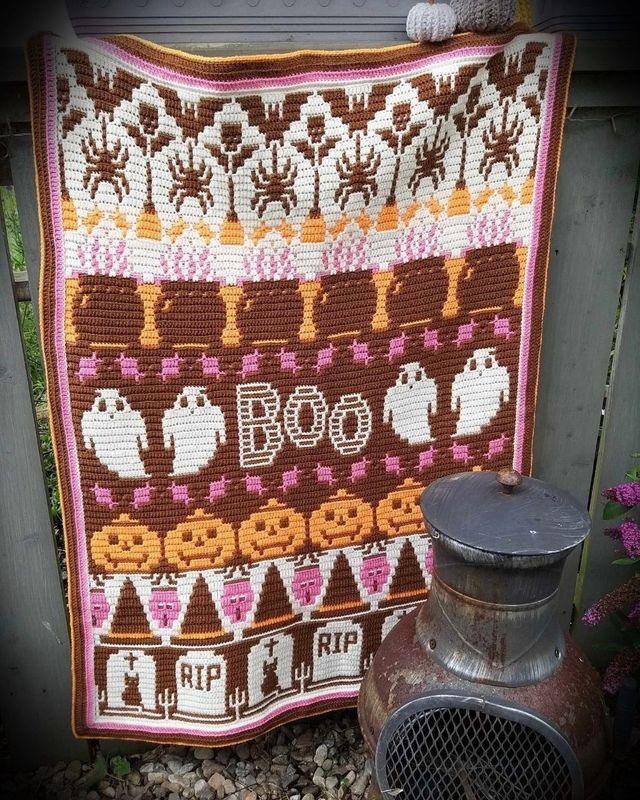 A crocheted blanket in cream, brown, orange and pink. The blanket contains various Halloween designs such as ghosts, pumpkins, spiders, zombies, cauldrons and more.