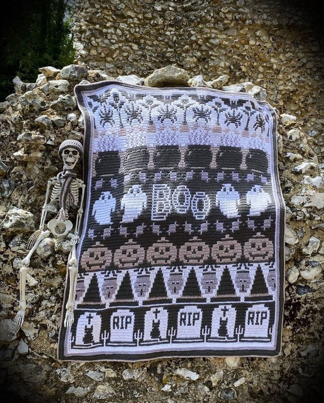 A crocheted blanket in white and grey. The blanket contains various Halloween designs such as ghosts, pumpkins, spiders, zombies, cauldrons and more.