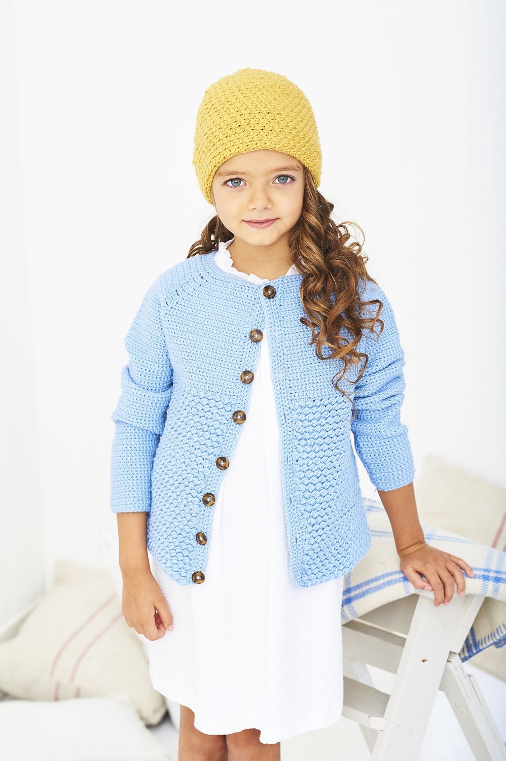A girl is wearing a blue crocheted cardigan and yellow crocheted hat.
