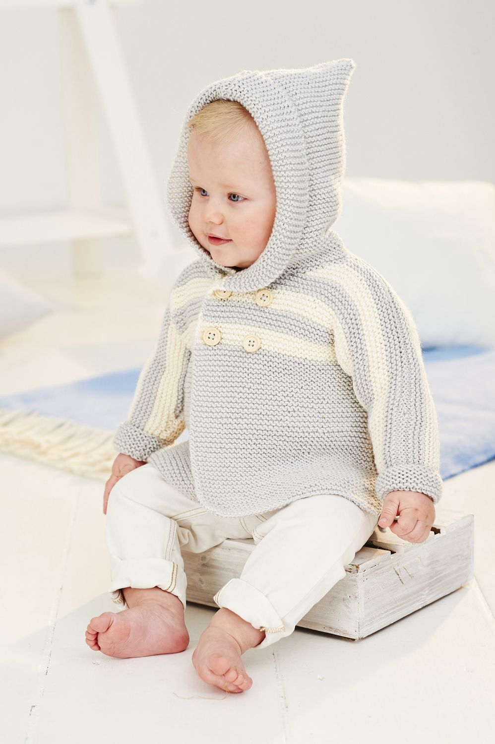 A baby is wearing a light grey knitted hooded cardigan with cream stripes.