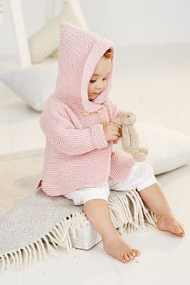 A baby is wearing a pale pink knitted hooded cardigan.