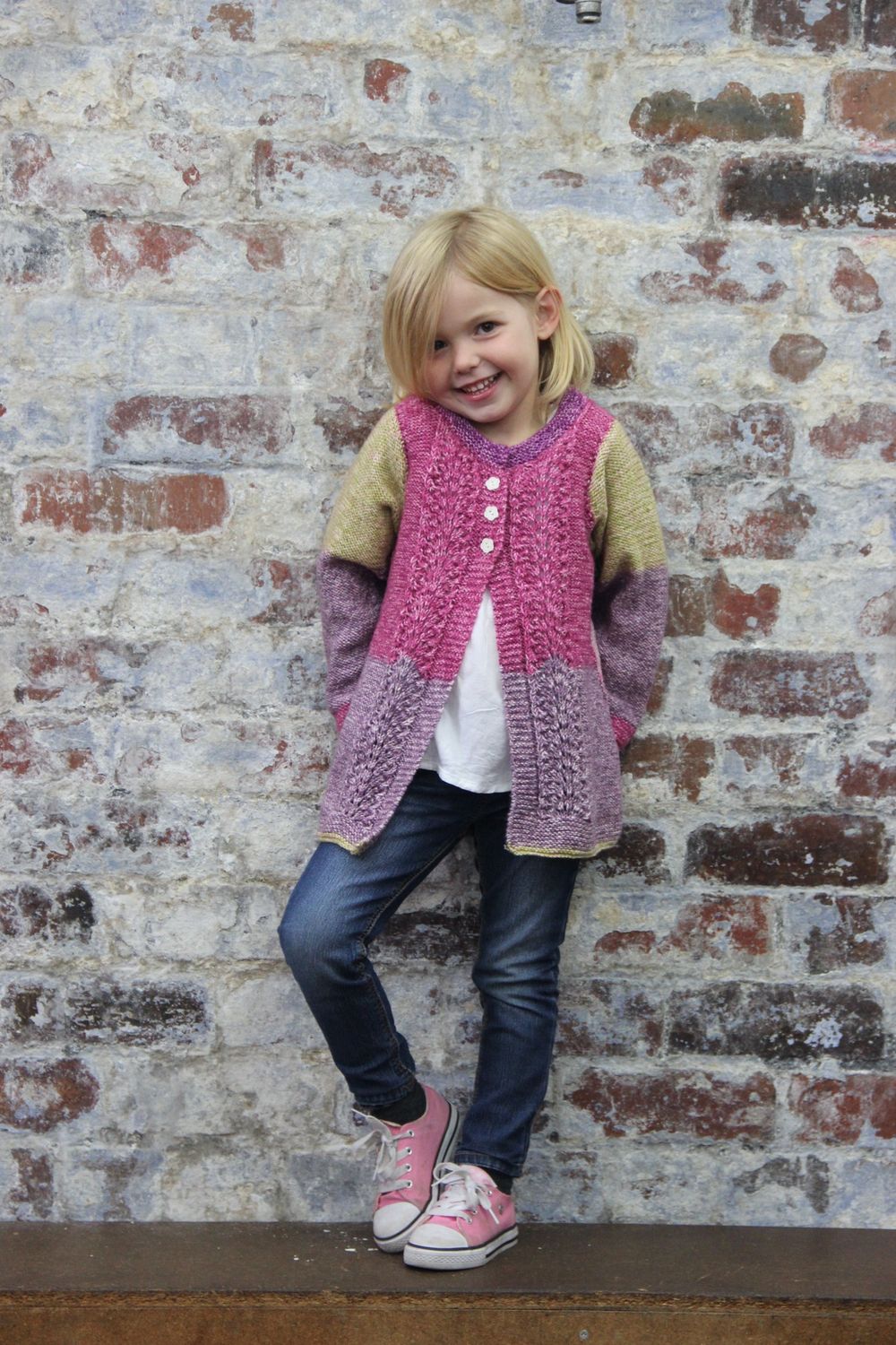 A girl is wearing a knitted cardigan in a variegated green, pink and purple yarn.