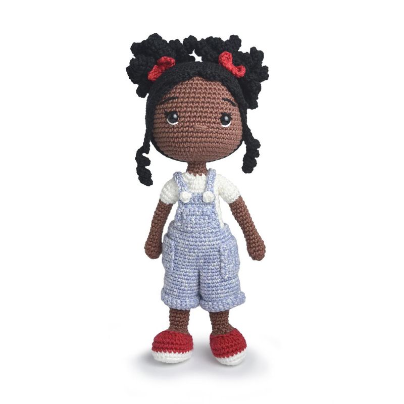 A crocheted doll, of a Black girl with black hair, a white t-shirt, blue dungarees and red shoes.